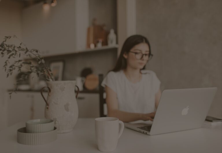 woman sitting at desk with Apple laptop open infront of her. On her desk in the foreground is a coffee cup, two bowls stacked on top of each other, and a vase with dried flowers inside.