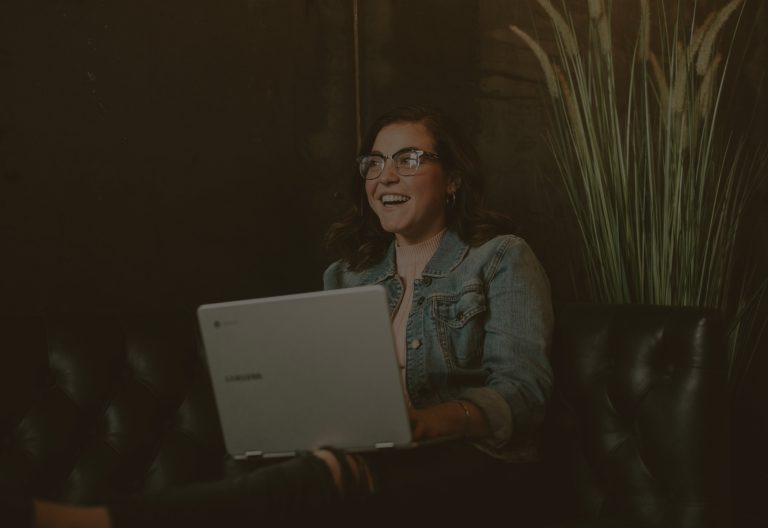 women on black leather couch with laptop on her lap, smiling and looking to her right. A tall bundle of grasses is behind the couch. Photo by Brooke Cagle on Unsplash.